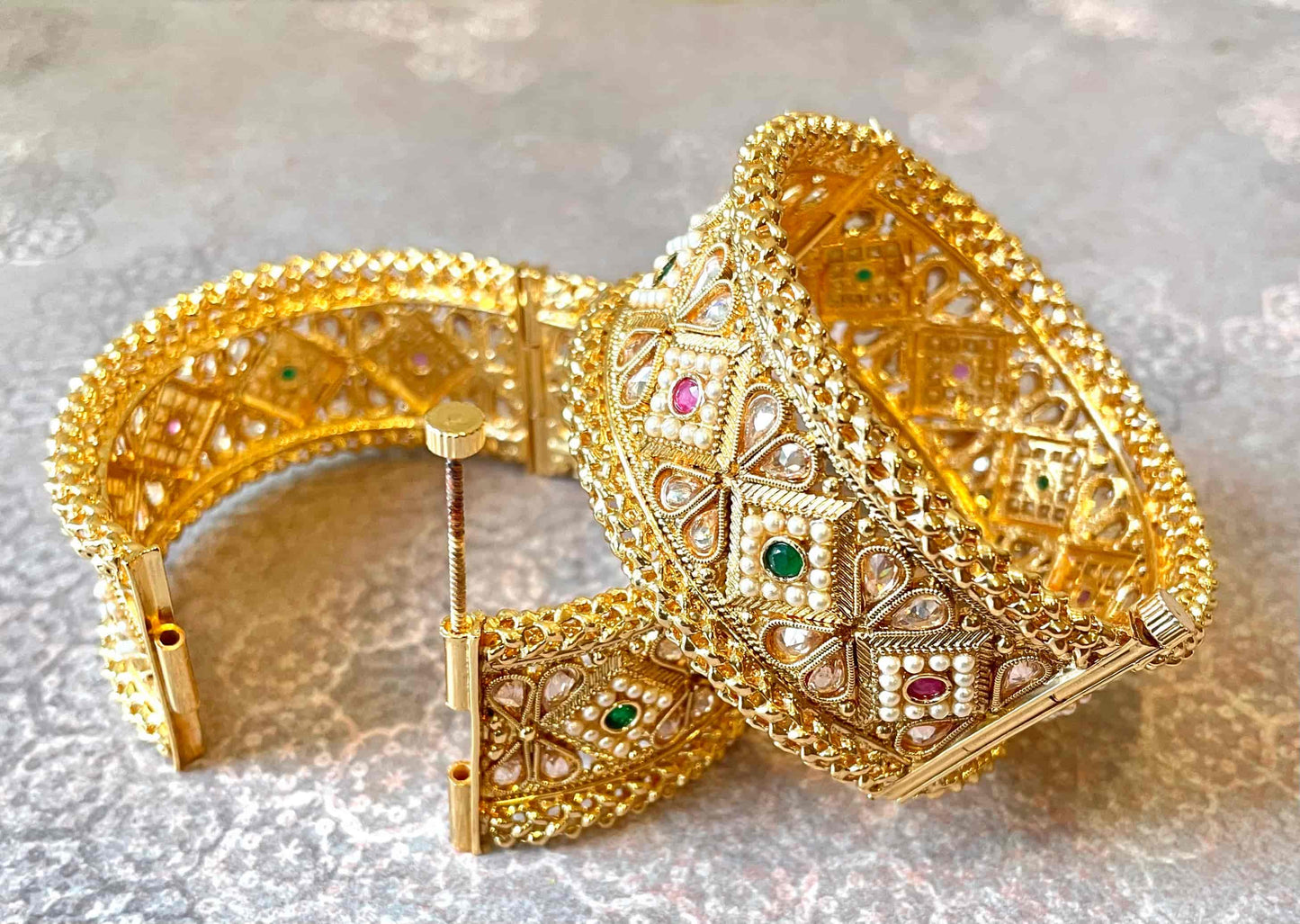 Two large-width gold bangles with intricate designs and gemstones on a light gray background