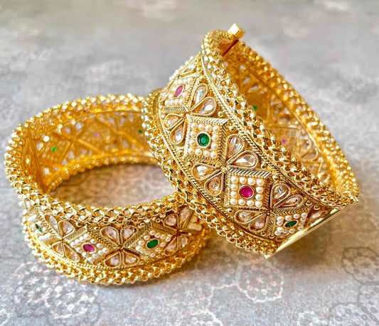 Two large-width gold bangles with intricate designs and gemstones on a light gray background
