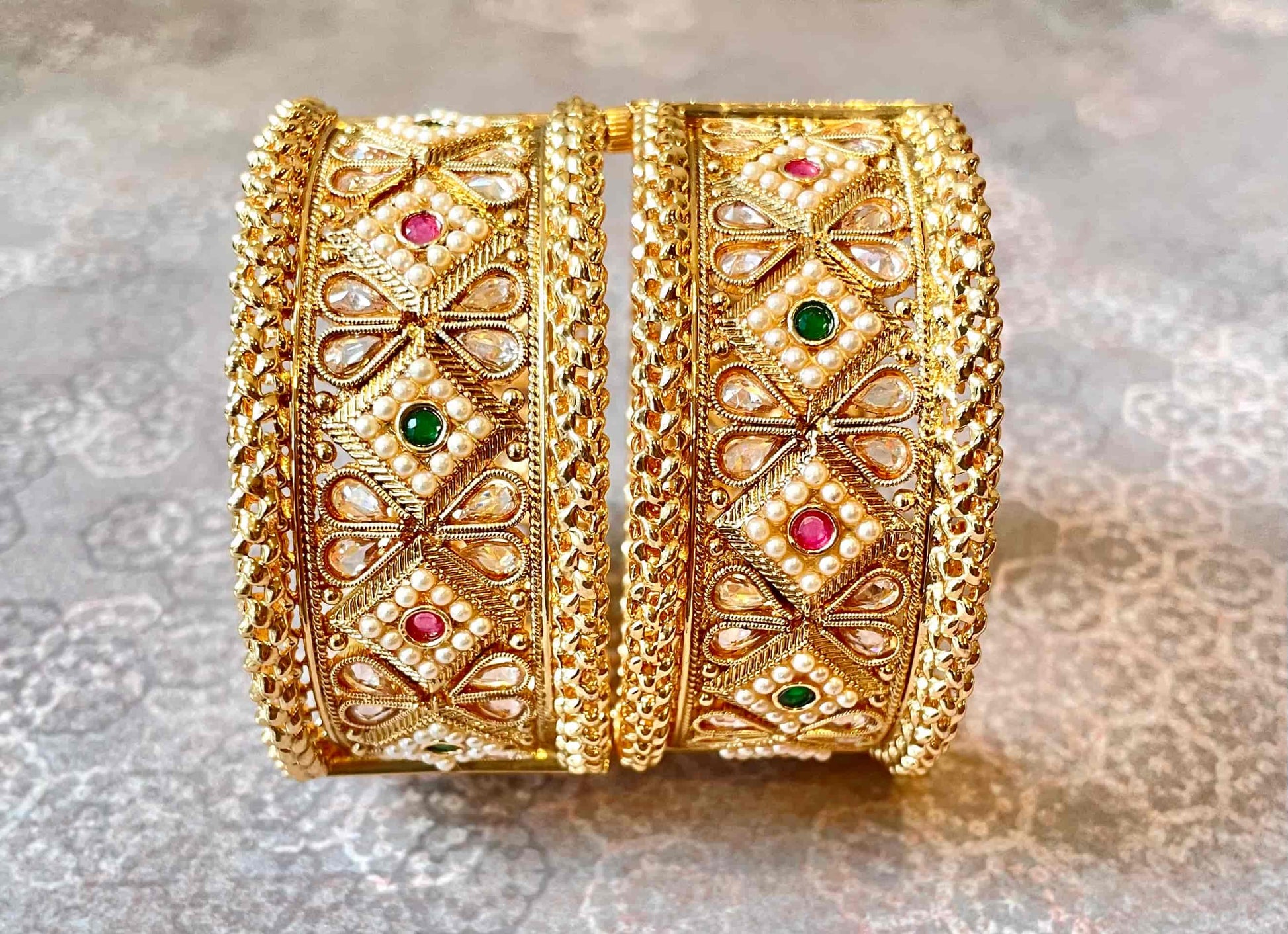 Two large-width gold bangles with intricate designs and gemstones on a light gray background