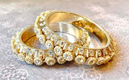 Two gold bangles with diamond accents and pearls surrounding them on a reflective surface