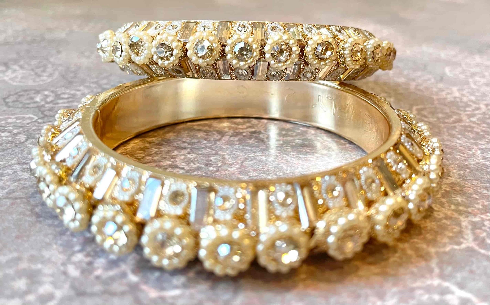 Two gold bangles with diamond accents and pearls surrounding them on a reflective surface