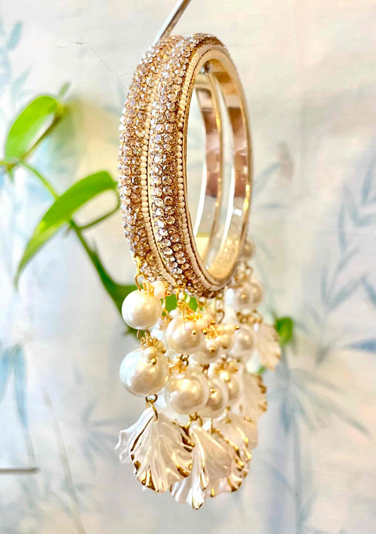 Sparkly golden bangles with hanging pearl and shell designs on a blurred natural background