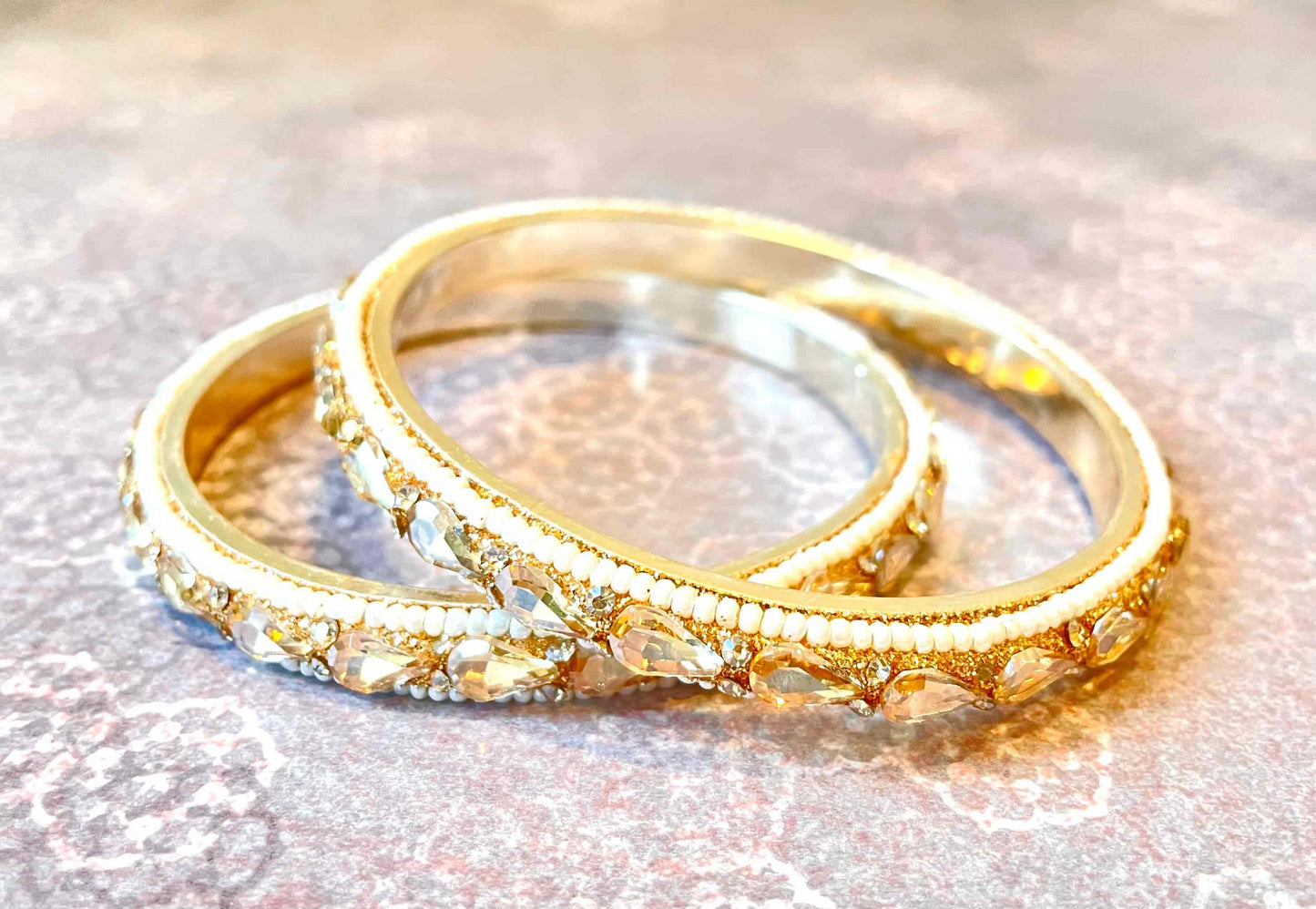 Two golden bangles with clear gemstones shaped as water droplets on a soft pink background
