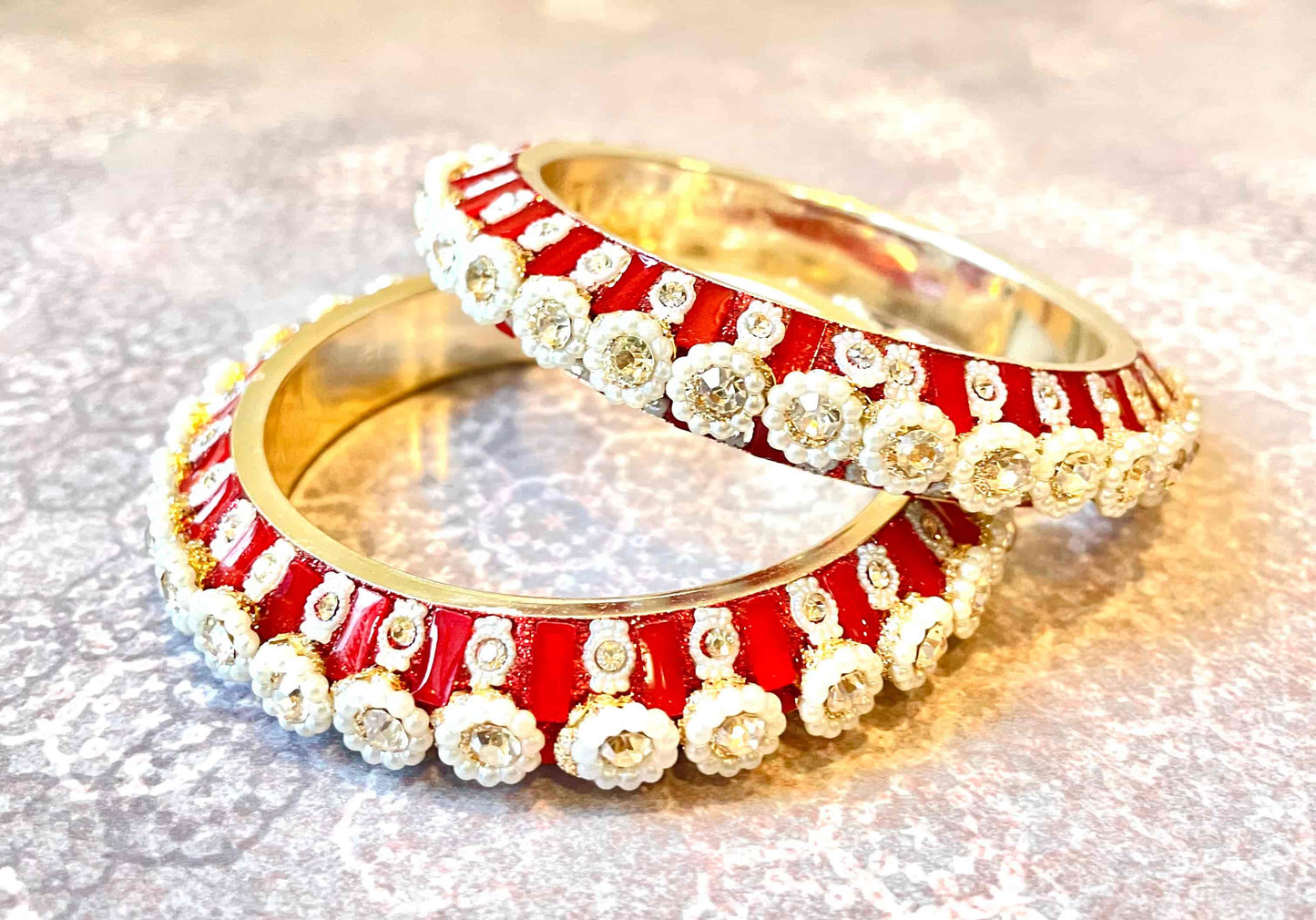 Two red and gold bangles with pearly floral designs and diamond accents on a reflective surface