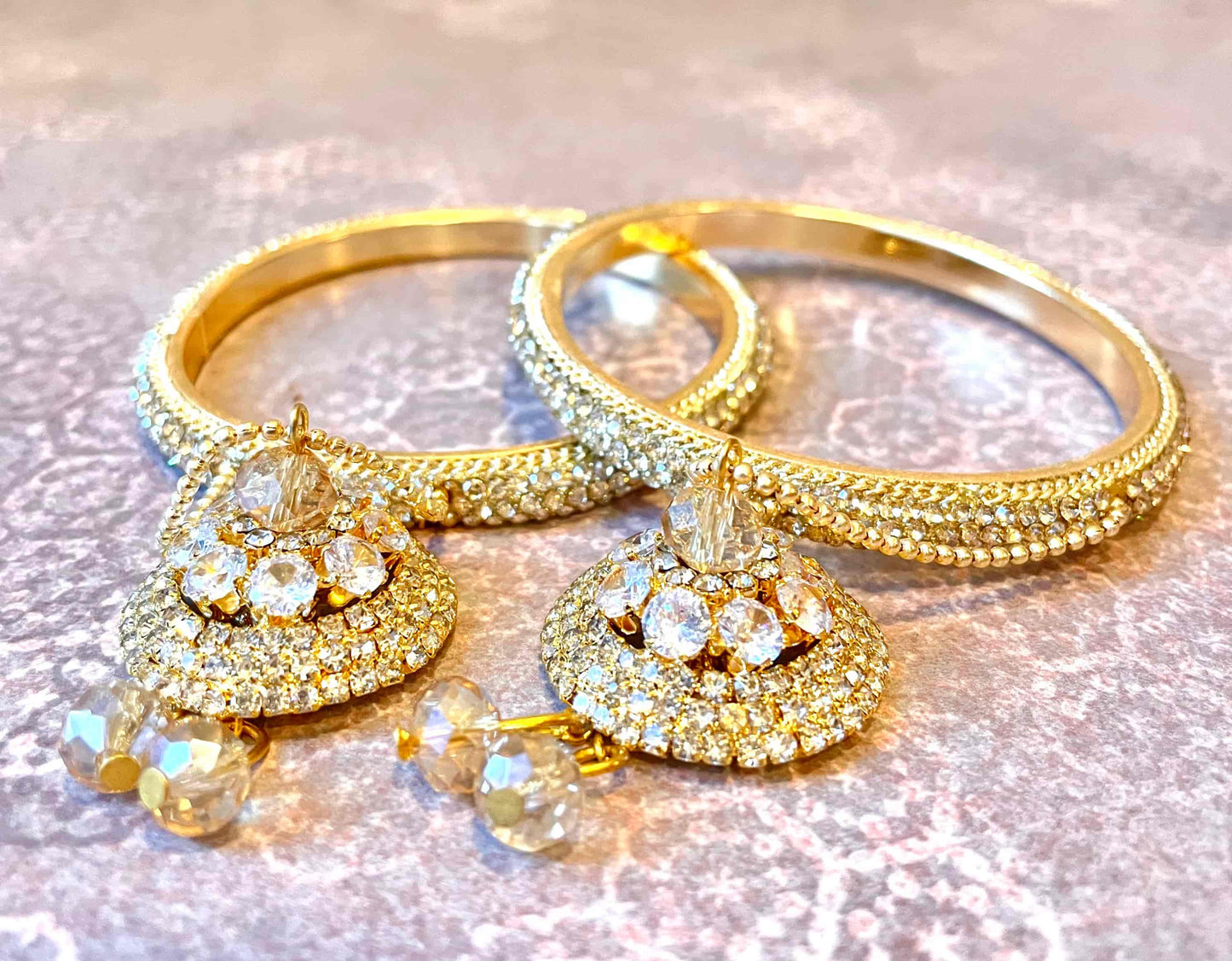 Sparkly pair of golden bangles with intricate designs and hanging pendants on a reflective surface