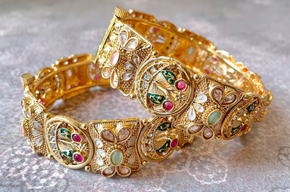 Gold bangles with gemstones and intricate peacock motifs on a textured gray background